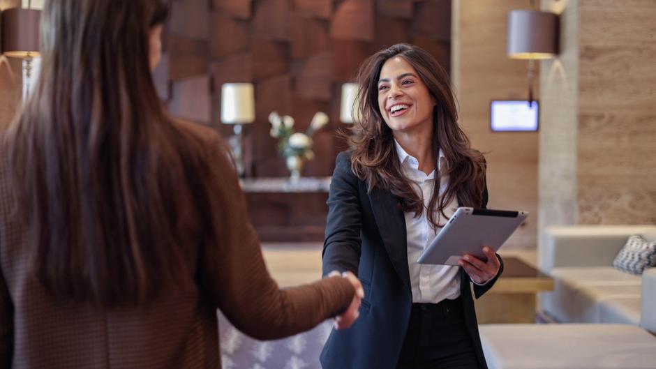 Zwei Frauen schütteln sich die Hände in einer modernen Lobby, eine hält ein Tablet.