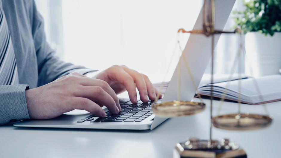 Hände tippen auf einer Laptop-Tastatur, im Vordergrund eine goldene Waage und ein offenes Notizbuch.