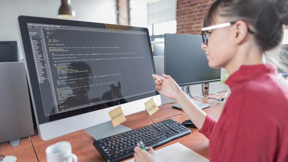 Eine Person sitzt an einem Schreibtisch und zeigt auf einen Computerbildschirm mit Code.