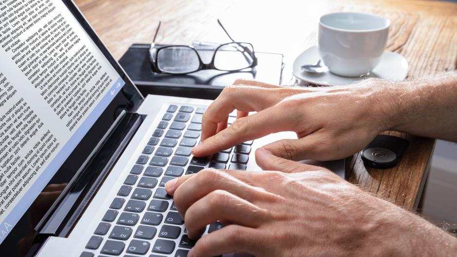 Hände tippen auf einer Laptop-Tastatur, im Hintergrund eine Tasse Kaffee und eine Brille.