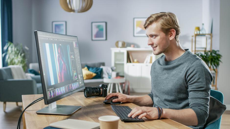 Ein junger Mann arbeitet an einem Computer, bearbeitet ein Bild und hat eine Kamera auf dem Tisch.