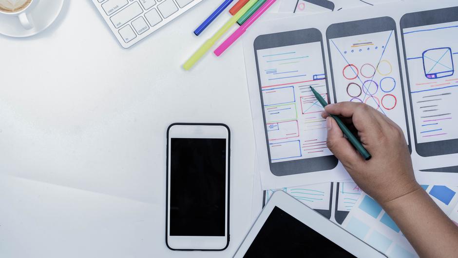 Auf einem Tisch liegen Skizzen von App-Layouts, ein Smartphone, ein Tablet, bunte Stifte und eine Tasse Kaffee.