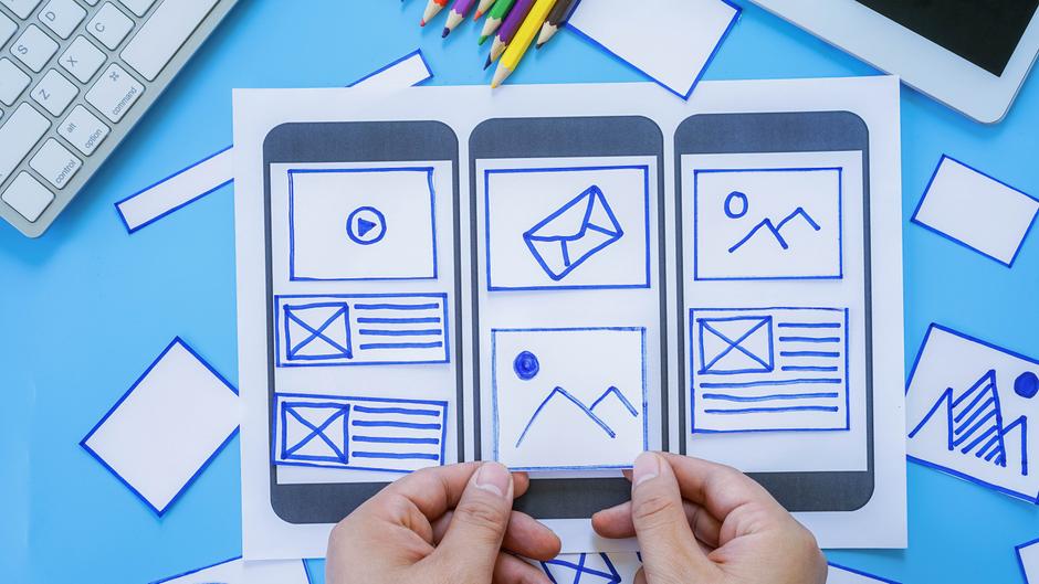 Hände halten ein Papier mit skizzierten Smartphone-Layouts, umgeben von bunten Stiften und weiteren Papierstücken.