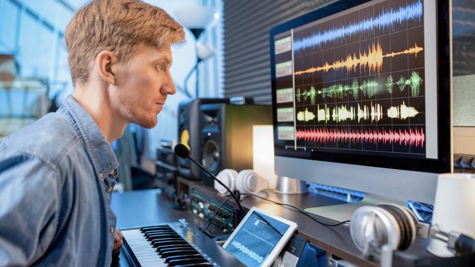 Mann sitzt an einem Schreibtisch mit einem Keyboard und einem Computer, auf dem Audio-Wellenformen angezeigt werden.