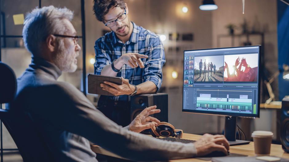 Zwei Personen arbeiten an einem Computer, einer zeigt auf ein Tablet, während der andere am Bildschirm mit Videobearbeitung beschäftigt ist.