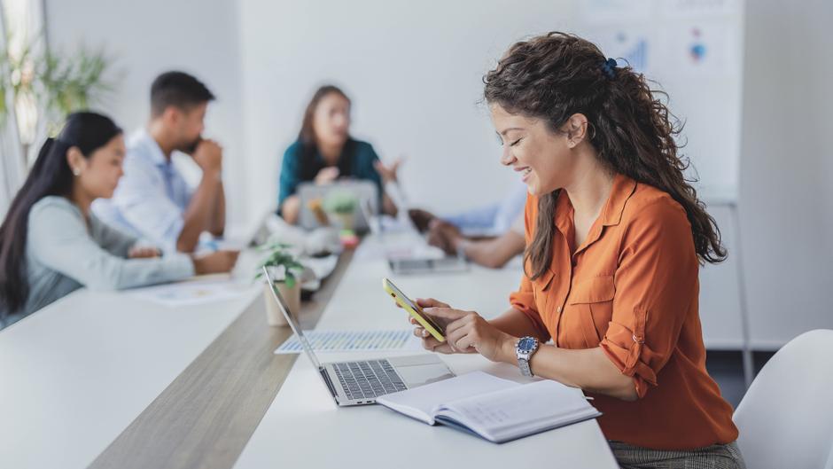 Eine lächelnde Frau in einem orangefarbenen Hemd nutzt ihr Smartphone an einem Tisch mit mehreren Personen und Laptops.