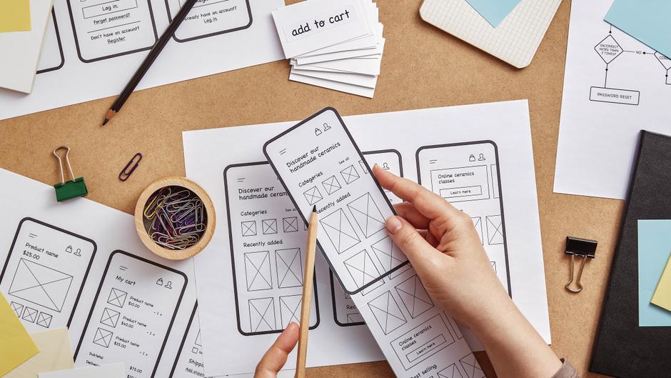 Skizzen von Smartphone-Layouts und Notizen auf einem Tisch, mit einer Hand, die ein Layout hält.
