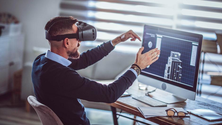 Mann mit VR-Brille interagiert mit einem Computerbildschirm, auf dem technische Zeichnungen angezeigt werden.