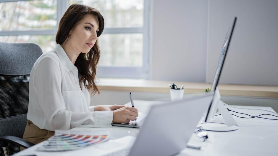 Frau mit langen Haaren sitzt an einem Schreibtisch, schreibt mit einem Stift und schaut auf einen Laptop.
