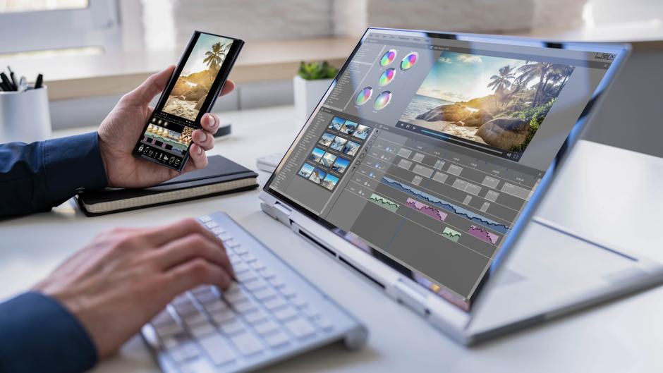 Eine Hand hält ein Smartphone, während die andere auf einer Laptop-Tastatur tippt; auf dem Bildschirm ist eine Bildbearbeitungssoftware sichtbar.