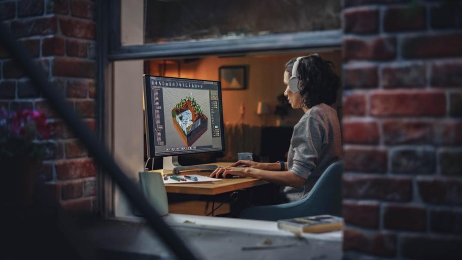 Eine Person mit Kopfhörern arbeitet an einem Computer, der ein 3D-Modell zeigt, in einem gemütlichen Raum.