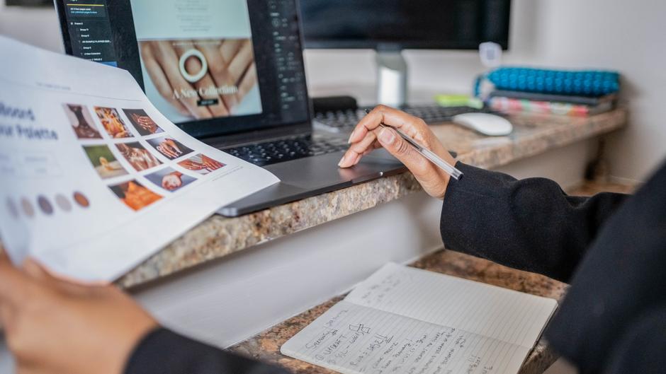 Eine Person hält ein Dokument mit Bildern in der linken Hand und tippt mit einem Stift auf einem Laptop.