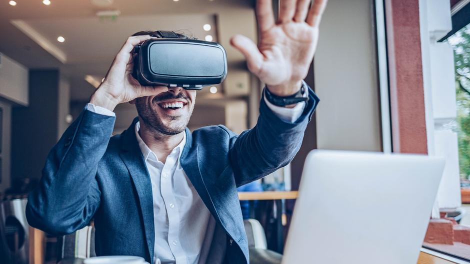 Mann mit VR-Brille lächelt und hebt die Hand in einem modernen Raum mit Laptop auf dem Tisch.