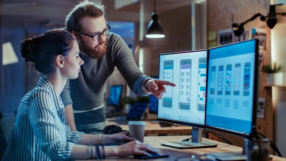 Zwei Personen arbeiten an einem Computer, während sie auf Smartphone-Designs auf dem Bildschirm zeigen.