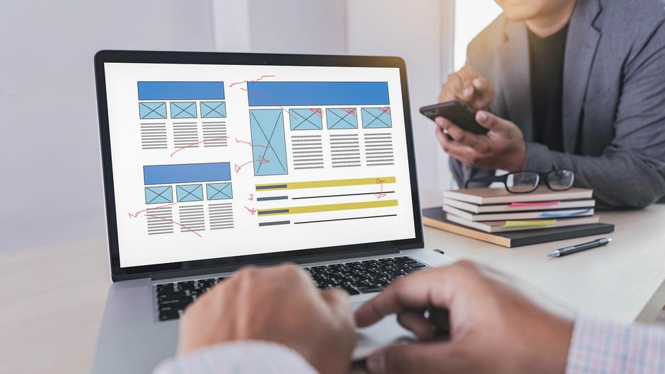 Auf dem Laptop-Bildschirm sind Wireframe-Layouts mit Anmerkungen zu sehen, während eine Person ein Smartphone hält.