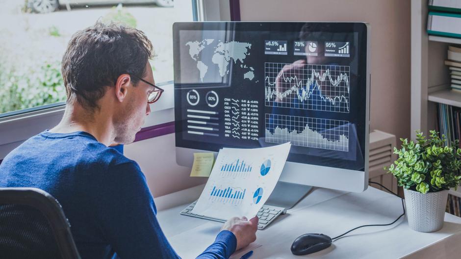 Mann mit Brille betrachtet Diagramme und Statistiken auf einem Computerbildschirm, während er Unterlagen in der Hand hält.