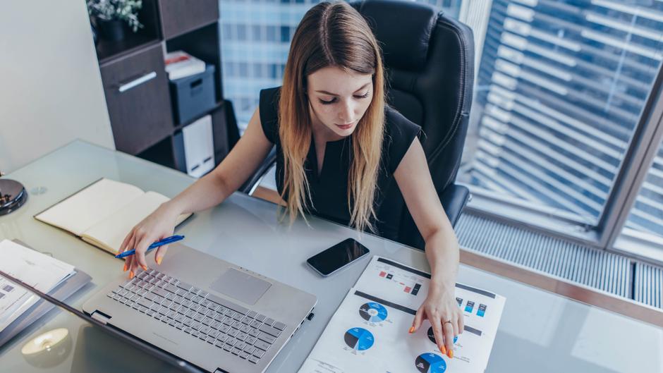 Eine Frau mit langen Haaren arbeitet an einem Schreibtisch mit Laptop, Notizbuch und Diagrammen.