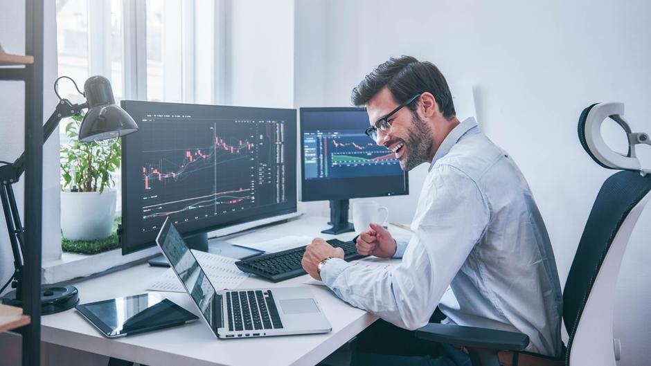Mann in einem modernen Büro lächelt und feiert vor mehreren Bildschirmen mit Finanzgrafiken.
