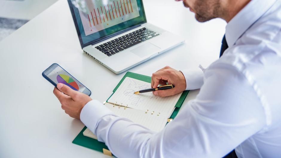 Mann in Hemd hält ein Smartphone und schreibt mit einem Stift in ein Notizbuch, Laptop und Diagramme im Hintergrund.