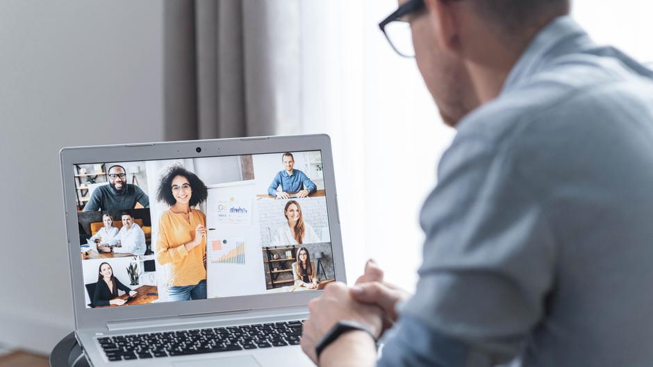 Mann sitzt vor einem Laptop, auf dem mehrere Personen in Videokonferenz zu sehen sind.