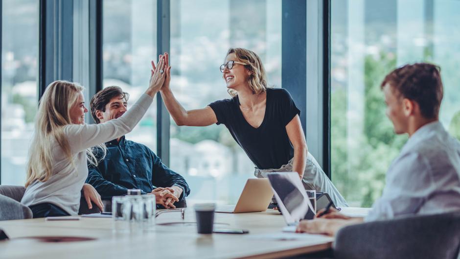 Zwei Personen geben sich in einem modernen Besprechungsraum ein High-Five, während zwei weitere Personen am Tisch sitzen.