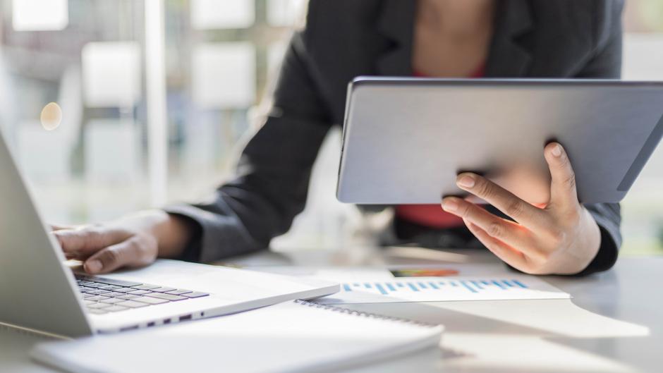 Eine Person hält ein Tablet in der Hand, während sie an einem Laptop arbeitet, umgeben von Notizen und Diagrammen.