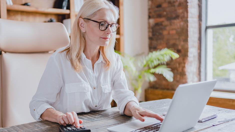 Eine Frau mit blonden Haaren und Brille arbeitet an einem Laptop und benutzt einen Taschenrechner.
