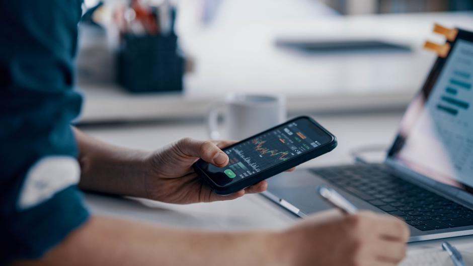 Eine Hand hält ein Smartphone mit einer Finanz-App, während eine andere Hand einen Stift über ein Laptop-Tastatur bewegt.