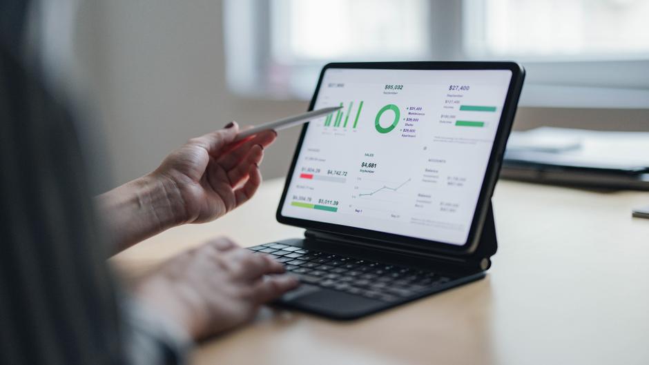 Eine Hand hält einen Stift und zeigt auf ein Tablet mit Finanzdaten und Diagrammen.