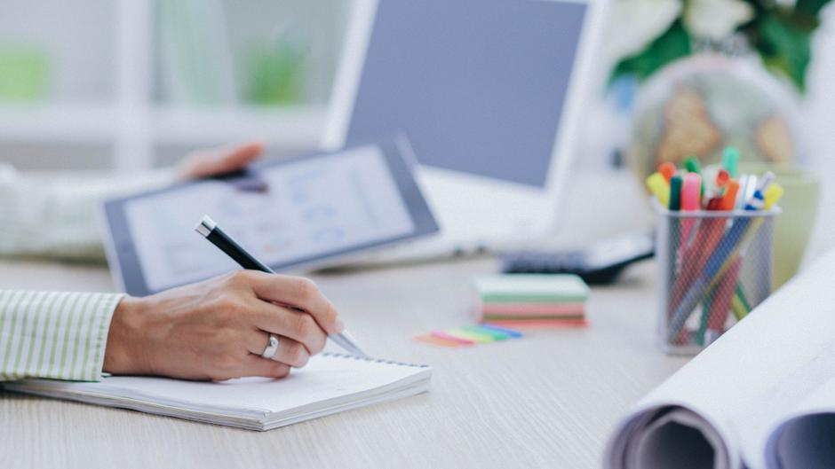 Eine Hand schreibt in ein Notizbuch, während ein Tablet und ein Stift auf dem Tisch liegen, umgeben von Schreibmaterialien.