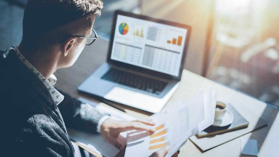 Mann mit Brille betrachtet Dokumente und Laptop mit Diagrammen und Tabellen auf einem Tisch.
