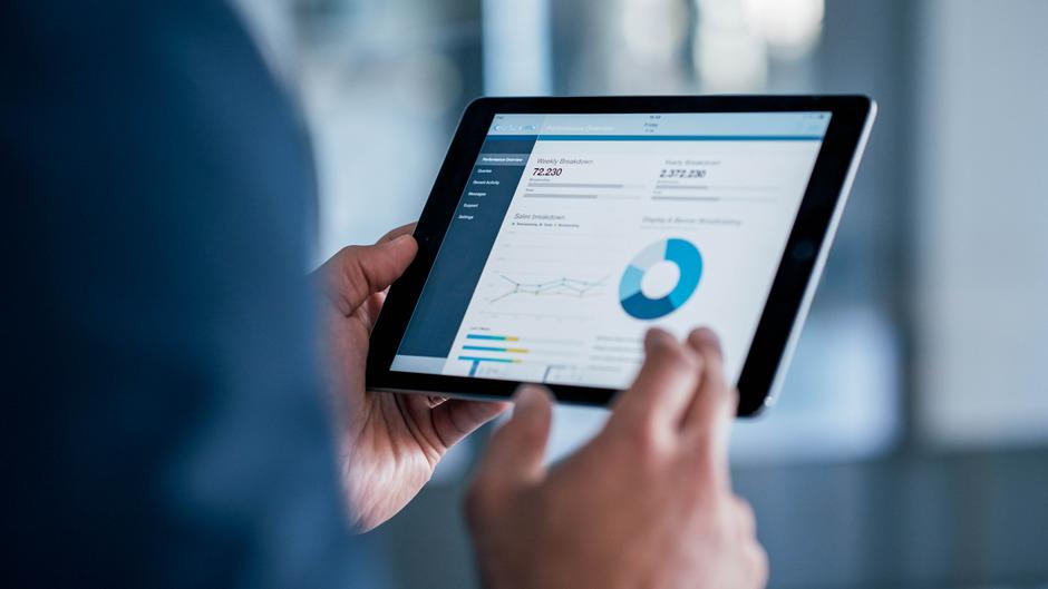 Eine Person hält ein Tablet, auf dem eine grafische Übersicht von Leistungsdaten angezeigt wird.