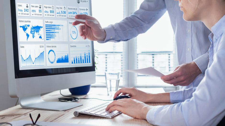 Zwei Personen analysieren Daten auf einem Computerbildschirm mit Diagrammen und Statistiken.