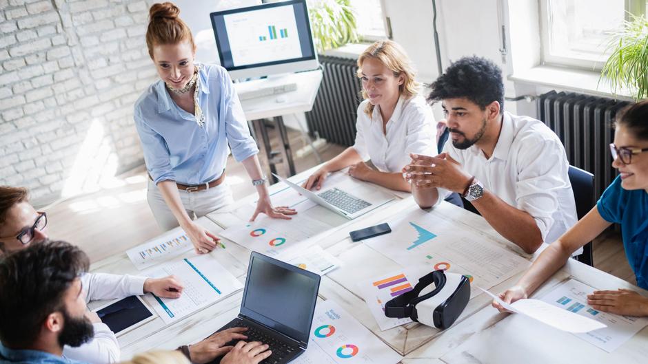 Eine Gruppe von sieben Personen diskutiert an einem Tisch mit Laptops und Diagrammen.
