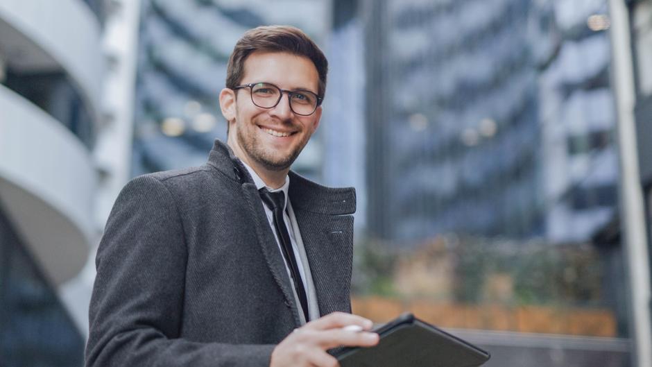 Lächelnder Mann in einem grauen Mantel hält ein Tablet in der Hand, vor modernen Gebäuden im Hintergrund.