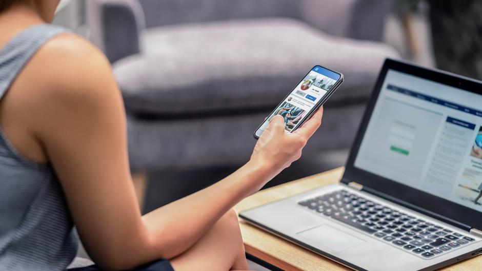 Eine Person hält ein Smartphone in der Hand und schaut auf den Bildschirm, während ein Laptop auf einem Tisch steht.