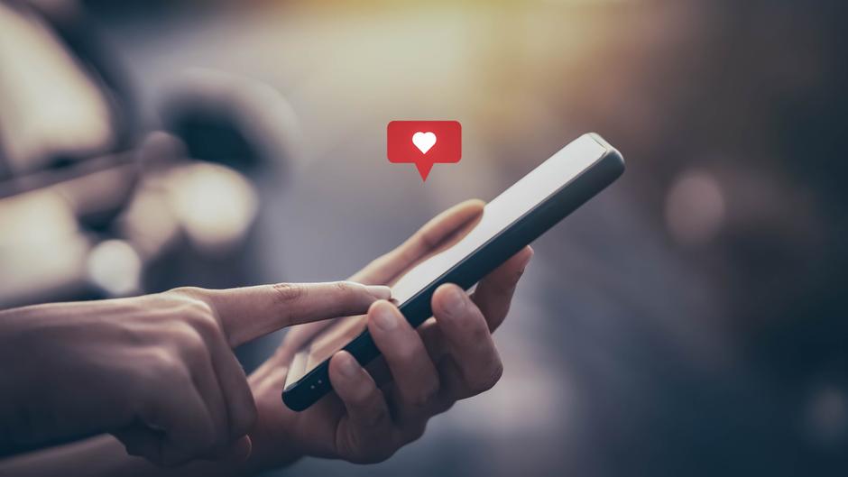 Eine Hand hält ein Smartphone, auf dem ein Herz-Symbol in einer Sprechblase angezeigt wird.