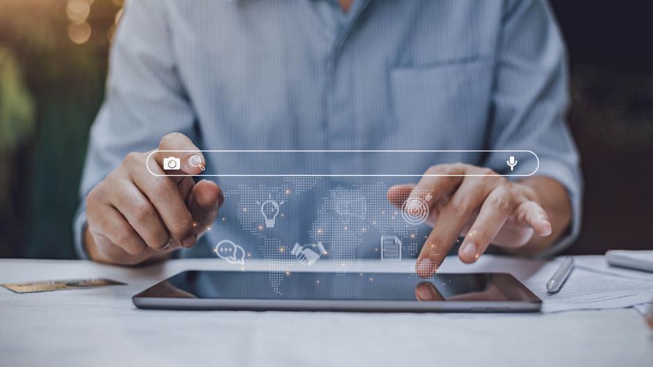 Hände tippen auf ein Tablet, während digitale Icons und eine Suchleiste über dem Bildschirm angezeigt werden.