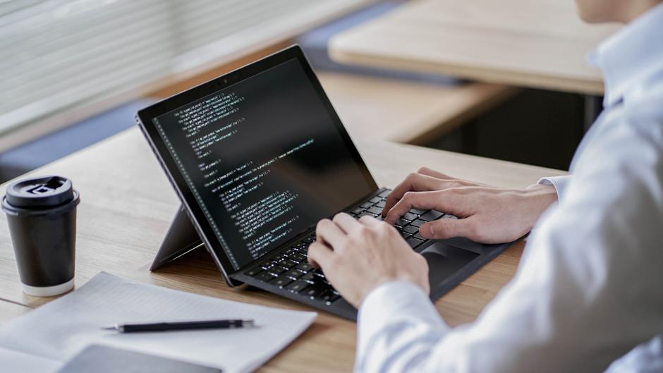 Eine Person tippt auf einem Laptop mit Code auf dem Bildschirm, daneben ein Kaffeebecher und ein Notizblock.