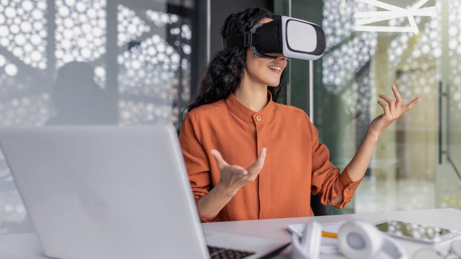 Frau mit VR-Brille sitzt an einem Tisch, gestikuliert und spricht, ein Laptop und Kopfhörer sind sichtbar.