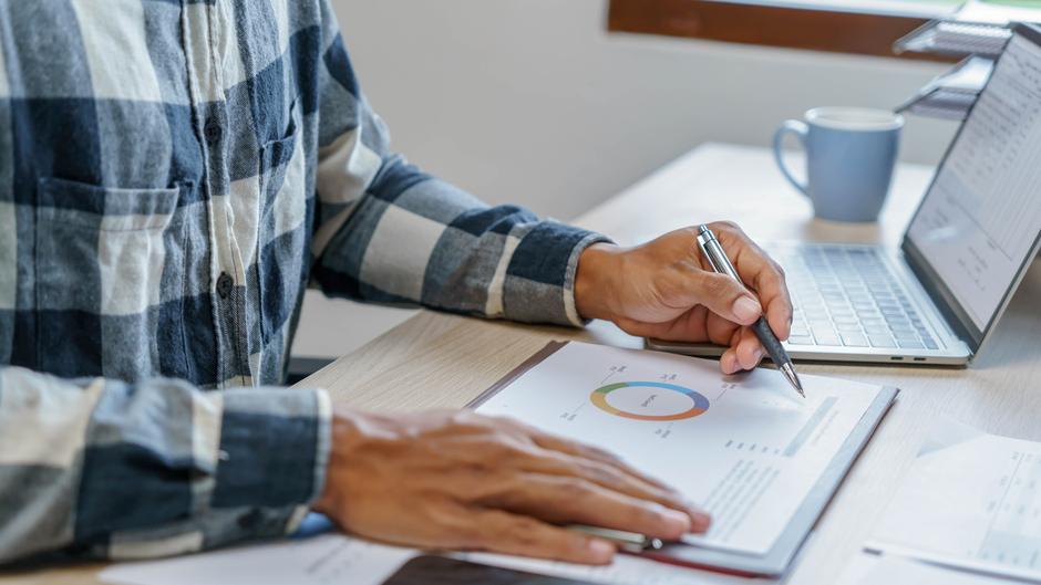 Hände eines Mannes, die einen Stift halten und auf ein Diagramm auf einem Blatt Papier zeigen, während ein Laptop und eine Tasse im Hintergrund sichtbar sind.