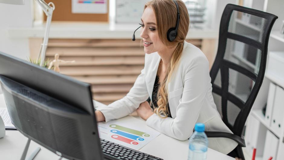 Frau mit Headset sitzt an einem Schreibtisch und arbeitet am Computer, neben ihr steht eine Wasserflasche.