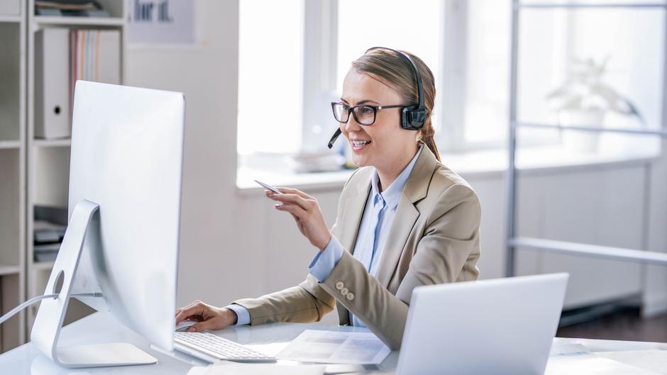 Frau mit Headset sitzt an einem Schreibtisch und spricht lächelnd in ein Mikrofon, während sie auf einen Computerbildschirm schaut.