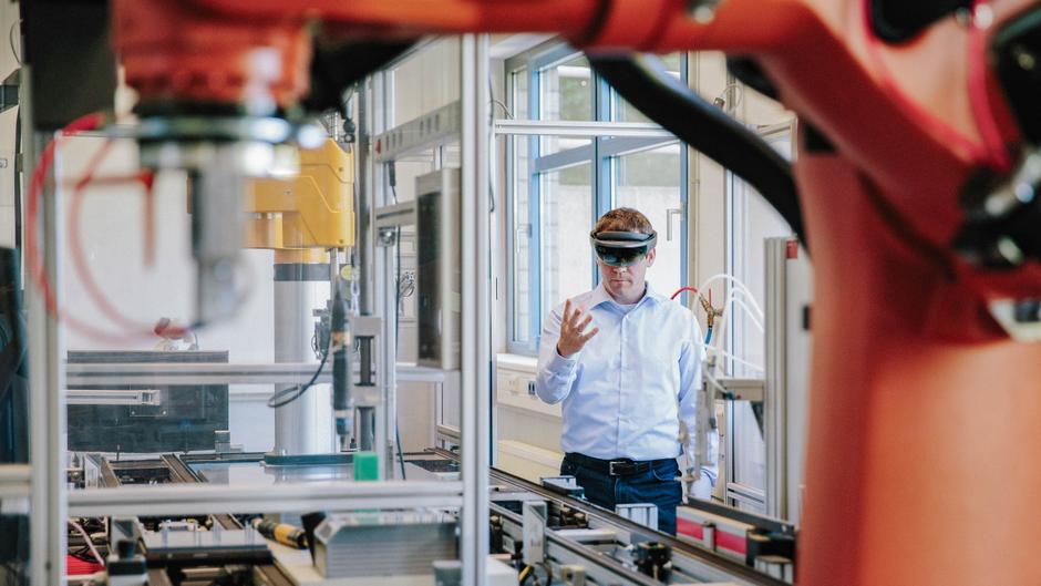 Ein Mann mit einer Datenbrille steht in einem technischen Labor neben einem Industrieroboter und interagiert mit Maschinen.