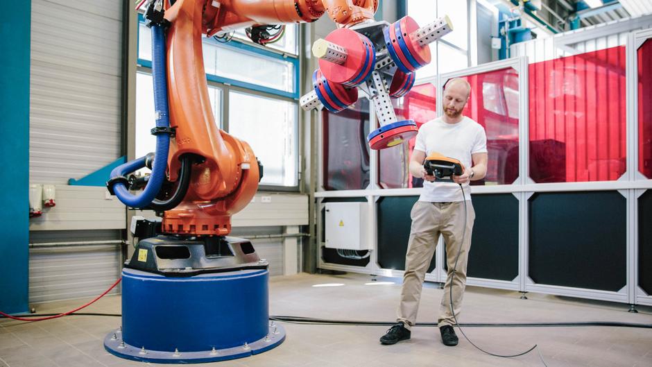 Ein Mensch steht vor einem Industrieroboter und hält ein Steuergerät in der Hand.