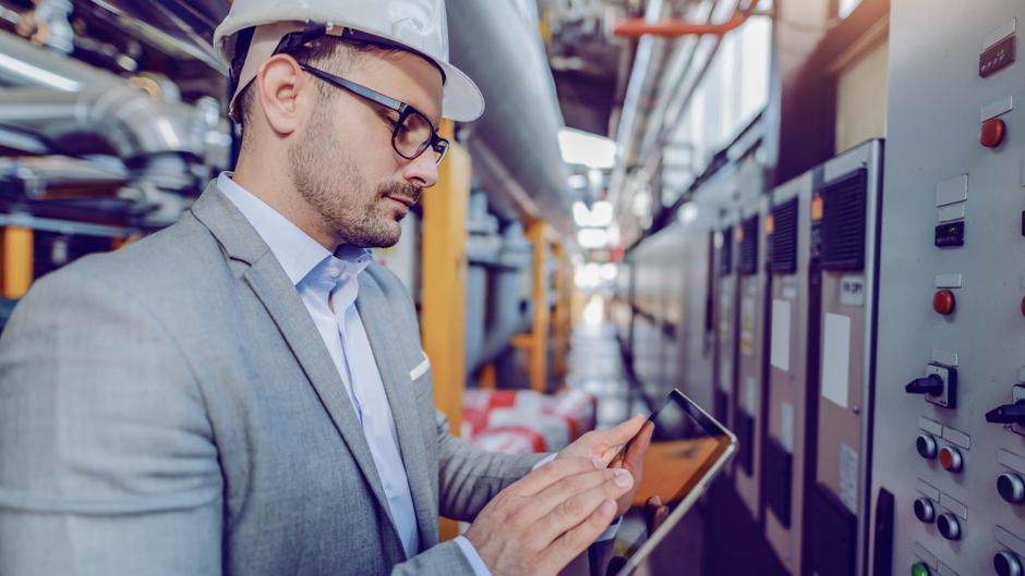 Mann in Anzug und Helm nutzt ein Tablet in einer industriellen Umgebung mit Maschinen und Steuerungstafeln.