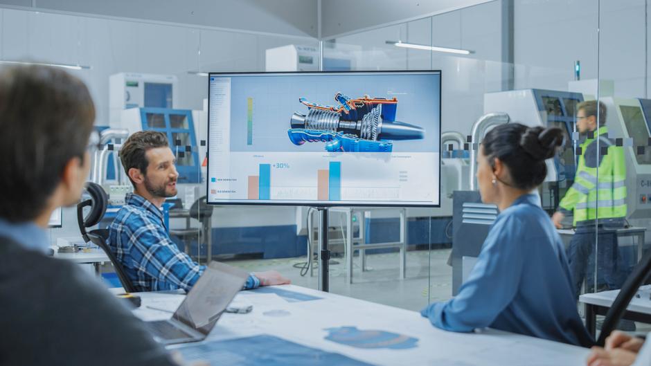 Eine Gruppe von Personen sitzt an einem Tisch und schaut auf einen Bildschirm mit technischen Grafiken und Diagrammen.