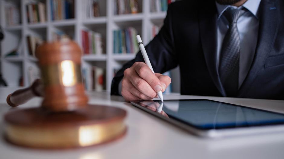 Ein Mann in Anzug schreibt mit einem Stylus auf einem Tablet, während ein Richterhammer im Vordergrund liegt.