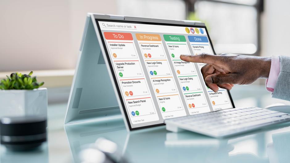 Ein Tablet mit einer Projektmanagement-Software zeigt Aufgaben in verschiedenen Status, während eine Hand darauf tippt.