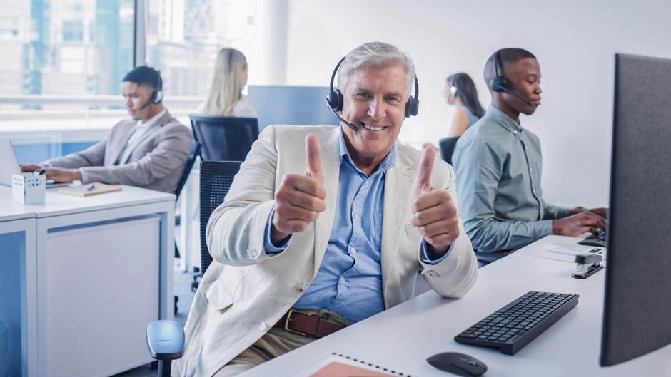 Ein älterer Mann mit Headset zeigt Daumen hoch, während er an einem Schreibtisch in einem Büro sitzt.
